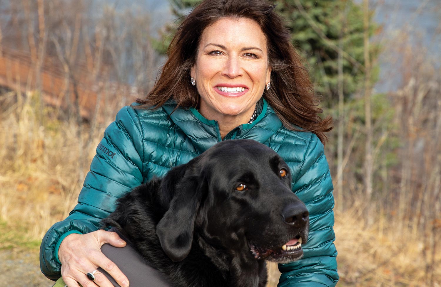 a woman smiles with her hands around a large black Labrador