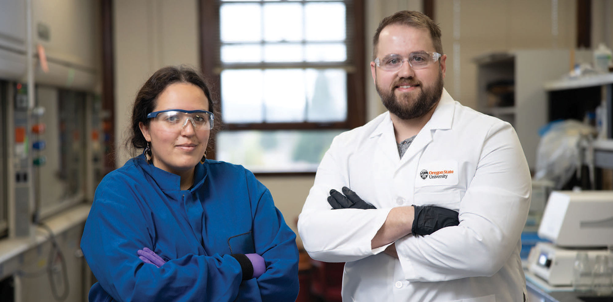 a woman wearing goggles and a blue jump suit stands beside a man wearing goggles and an Oregon State University lab coat, both with their arms folded and smiling while standing in a lab